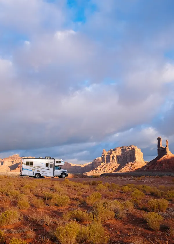 RV in golden scrubland below the red buttes of Valley of the Gods, with towering clouds.