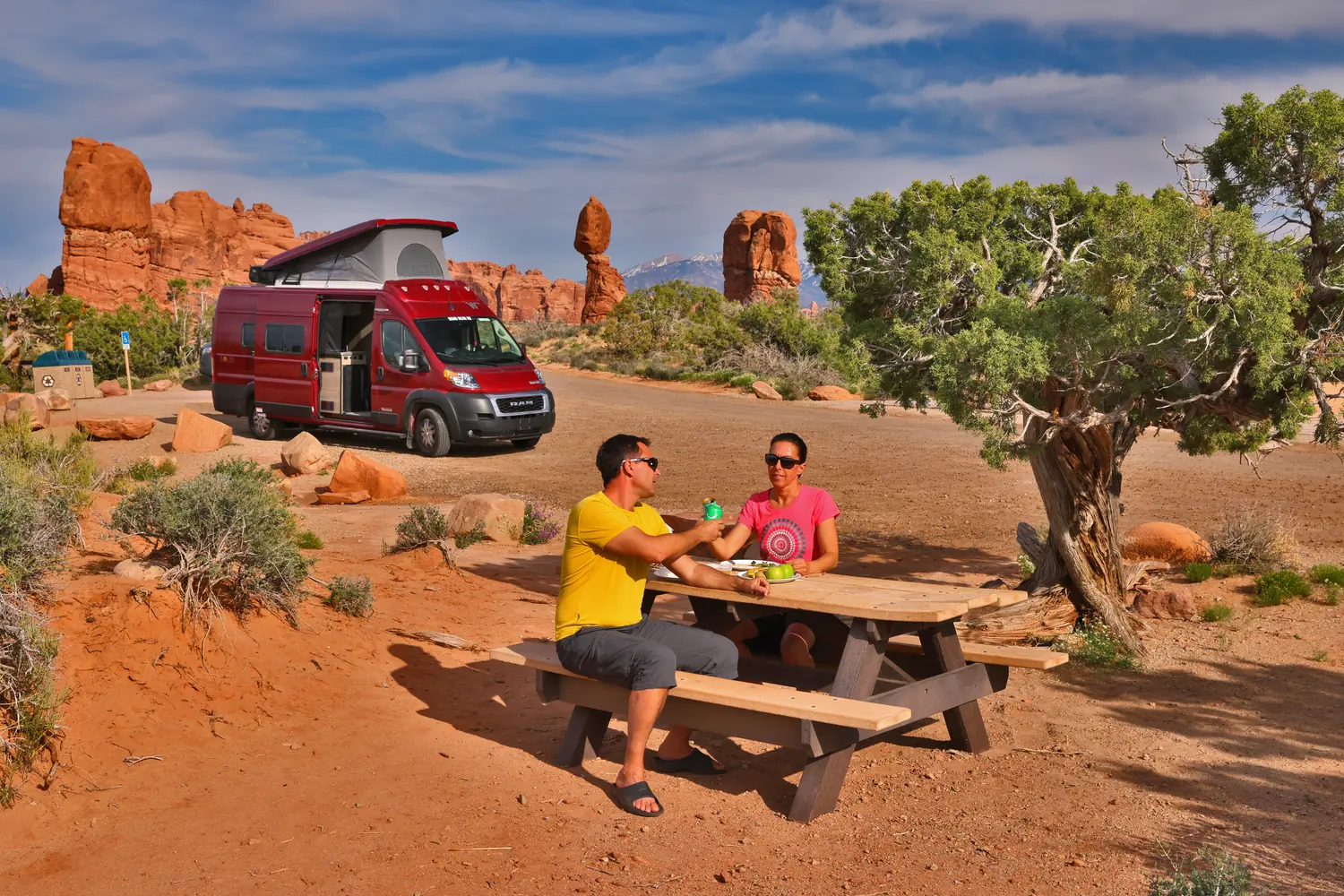 Smiling couple having a picnic next to their pop-top campervan among red rock spires
