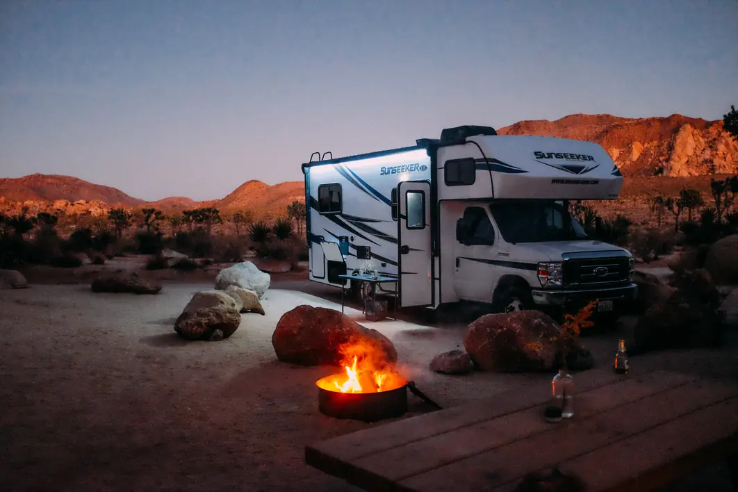 Campfire glowing beside a Class C motorhome at dusk among desert boulders