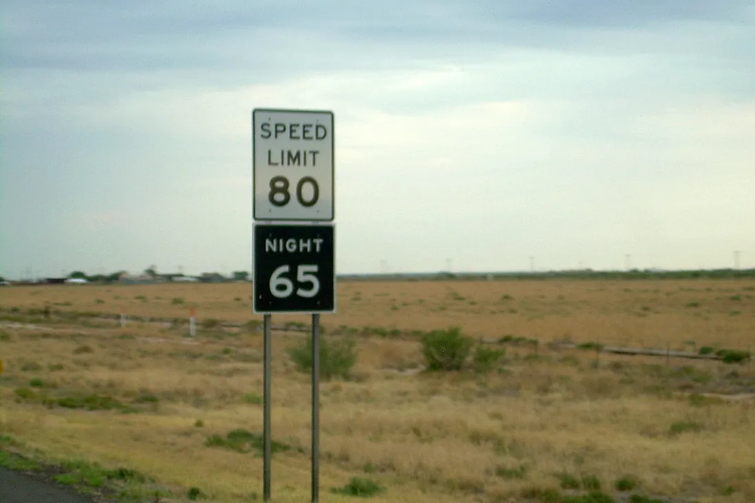 Texas speed limit sign: 80 mph by day and 65 mph at night, beside a flat grassland