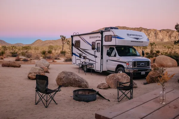 RV at a Joshua Tree campsite with camping chairs and a fire ring, at pink dusk