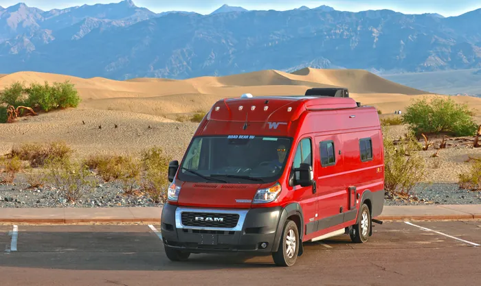 Red campervan parked in front of sand dunes and bare desert mountains