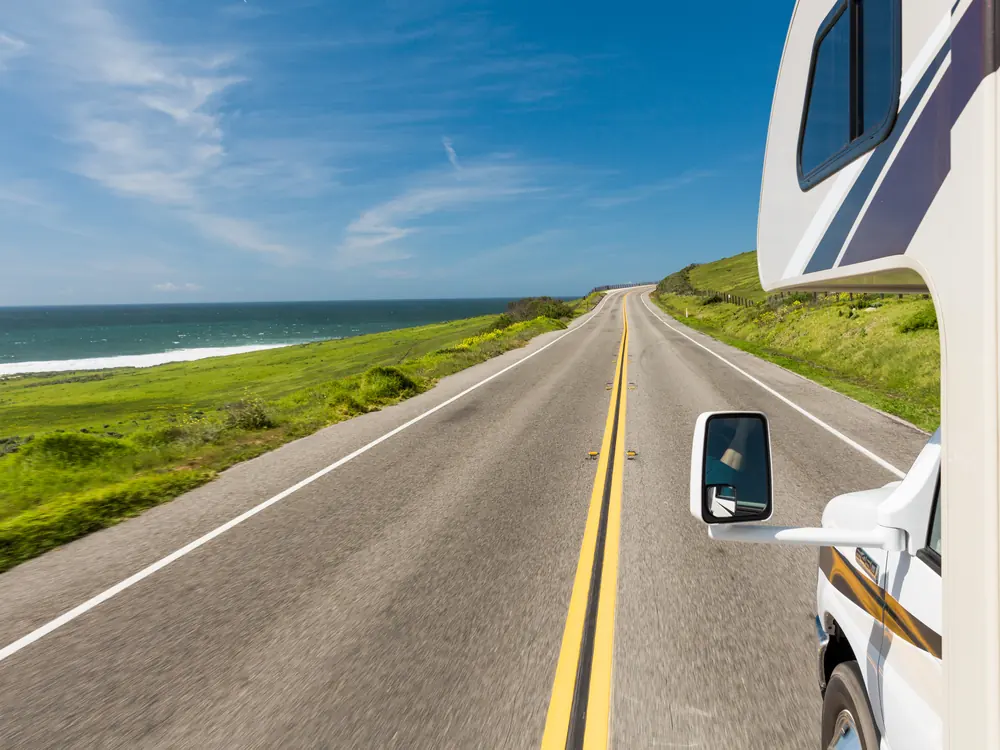 Straight coastal highway seen past an RV's side mirror, with green bluffs and the ocean