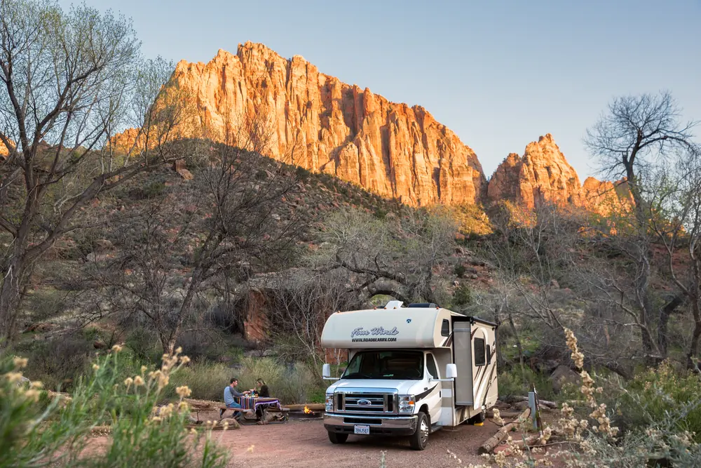 Couple eating at a picnic table next to their RV at a Zion campsite, with the Watchman glowing at sunset