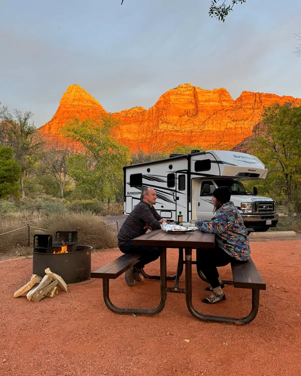 Couple at a picnic table beside their RV as the sunset lights up the red cliffs of Zion