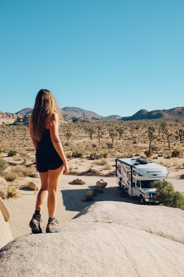 Woman standing on a granite boulder looking out over the Joshua Tree desert, with an RV parked below