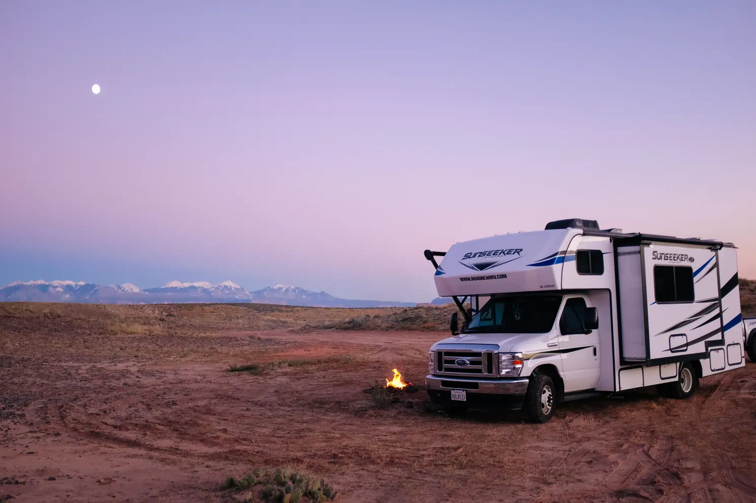 RV parked in the open desert at dusk next to a small campfire, under a pastel sky