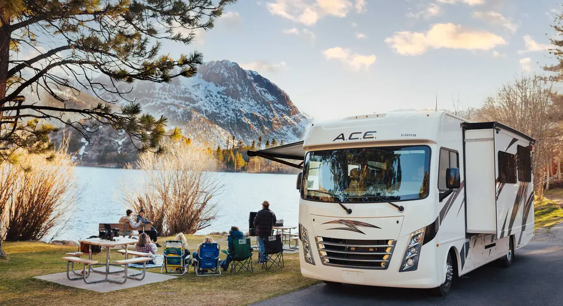 Motorhome at a lakeside campsite, with people at picnic tables and a snowy peak behind
