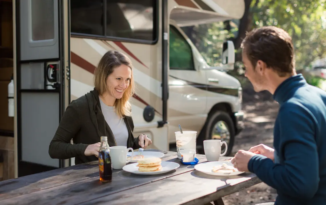 Smiling couple eating breakfast at a picnic table next to their RV